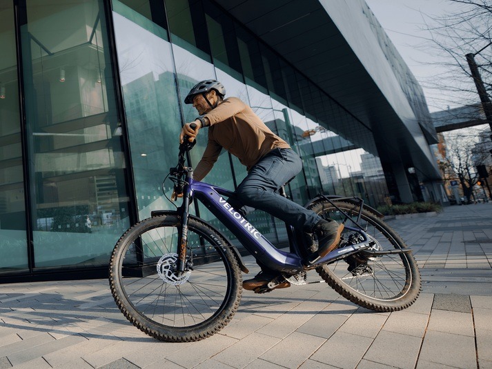 Helmeted rider on a Galaxy Purple Velotric Summit 2 outside a glass-fronted building Helmeted rider on a Galaxy Purple Velotric Summit 2 outside a glass-fronted building