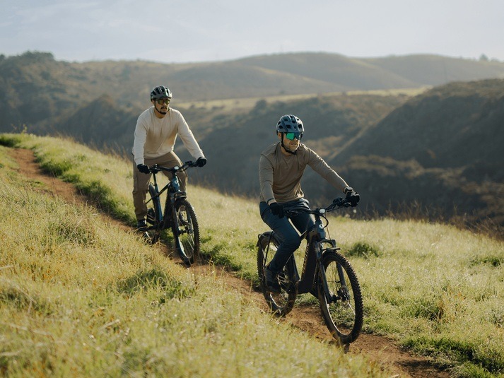 Two helmeted riders on Velotric Summit 2 bikes on a grassy track Two helmeted riders on Velotric Summit 2 bikes on a grassy track